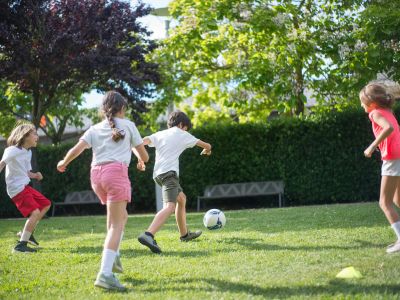 Kids playing soccer in a park with lush greenery and sunny weather.